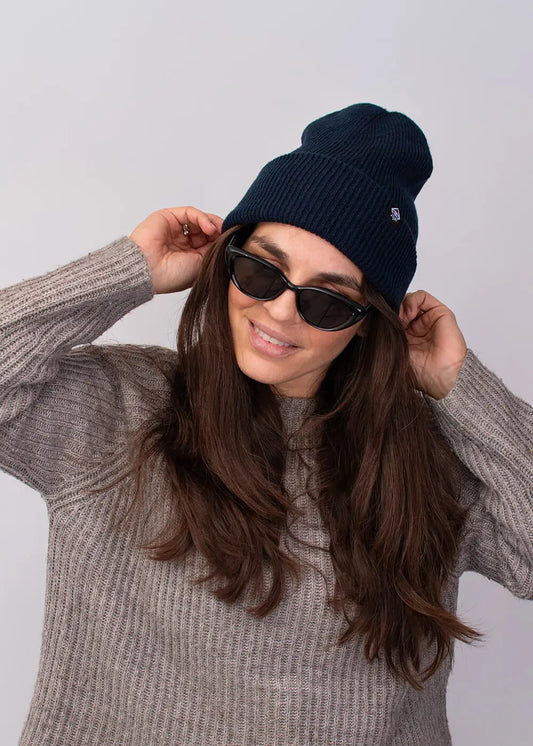 Eine Frau mit langen braunen Haaren trägt die J.Clay Deep Cuffed Beanie French Navy, eine dunkle Sonnenbrille und einen grauen Strickpullover. Sie lächelt und rückt ihre Mütze mit beiden Händen vor einem schlichten hellen Hintergrund zurecht.