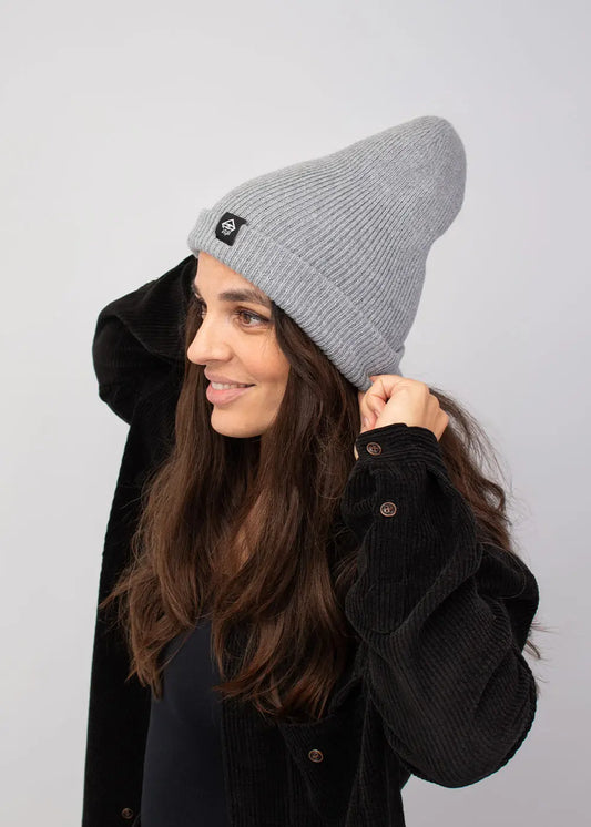 Eine Frau mit einer J.Clay Deep Cuffed Beanie Ash und einer schwarzen Jacke lächelt und rückt ihren Hut zurecht.