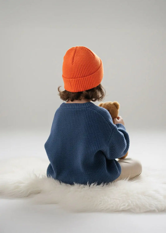 Ein Kind mit einer J.Clay Kids Beanie Sunset und einem blauen Pullover sitzt auf einem weißen Teppich und hält einen Teddybären.