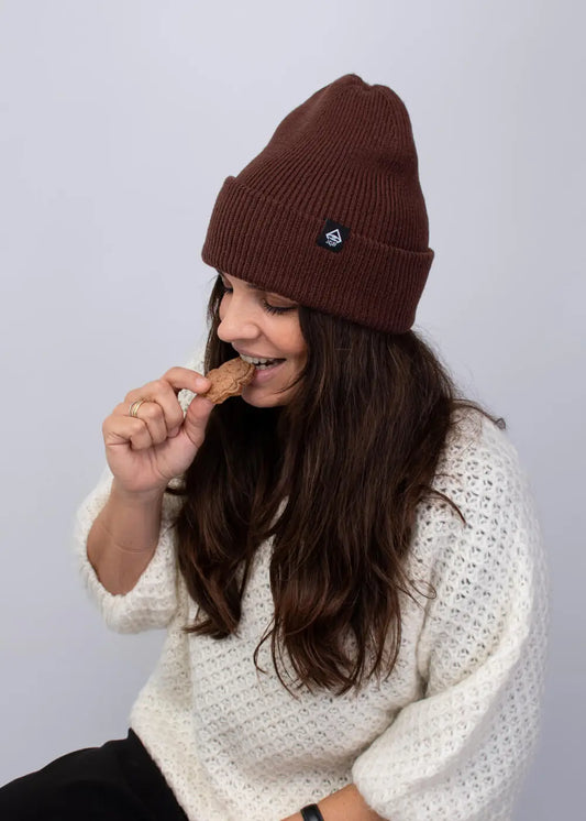 Frau in einem J.Clay Deep Cuffed Beanie Walnut und weißem Pullover isst einen Keks.