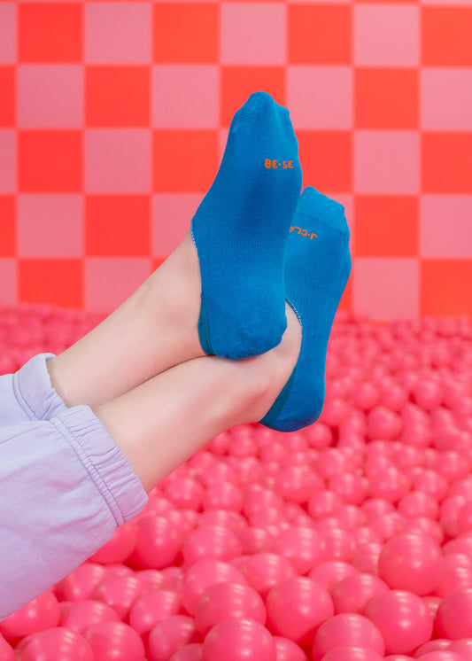 Zwei Füße in hellblauen J.Clay Füßlingen, die nicht rutschen, stehen vor rosa Bällen und einer rot-rosa karierten Wand. Die Trägerin, gekleidet in helllila Hosen, genießt den Komfort der J.Clay unsichtbaren Füßlinge.