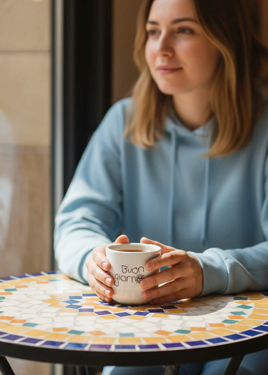 Frau im blauen Kapuzenpulli hält eine J.Clay Buongiorno - Tasse an einem bunten Mosaiktisch.