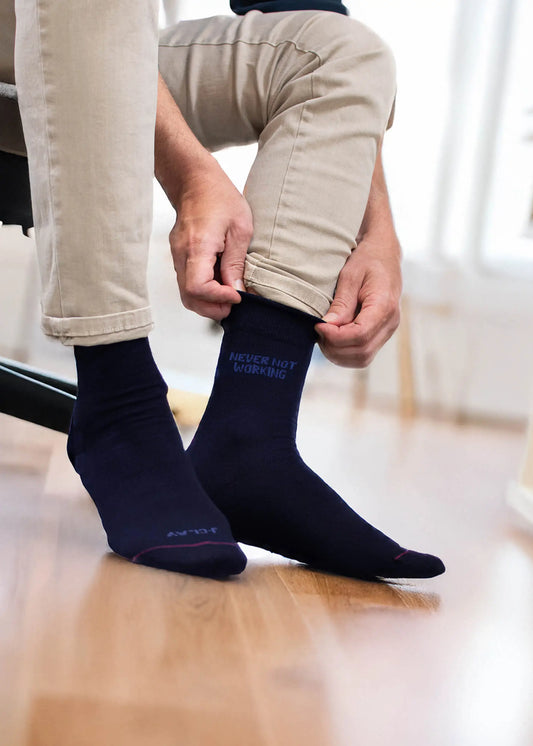Eine Person passt ihre marineblauen J.Clay-Business-Socken mit der Aufschrift "NEVER NOT WORKING" an, während sie auf einem Stuhl in einem hellen Raum sitzt, eine beigefarbene Hose trägt und einen Holzboden unter sich hat.