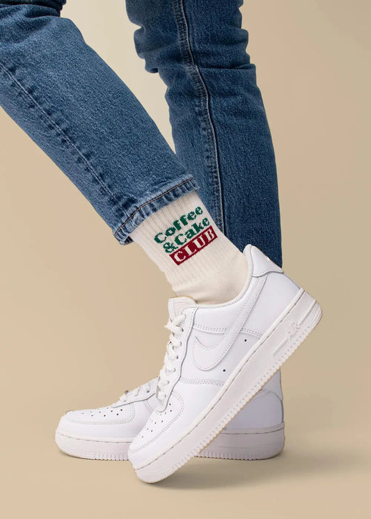 Person in blauen Jeans und weißen Turnschuhen, mit J.Clay Coffee Cake Club Socken.