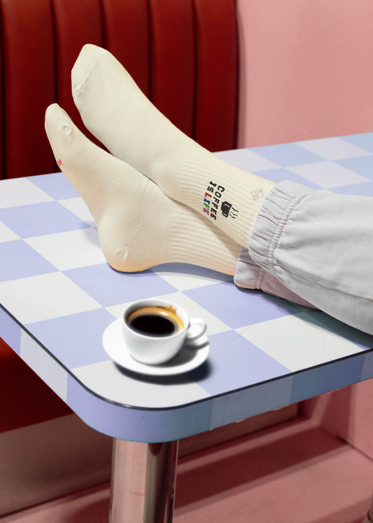 Eine Person in weißen J.Clay-Tennissocken mit Text ruht ihre Füße auf einem blau-weiß karierten Tisch aus, mit einer Tasse schwarzen Kaffees in der Nähe vor einem rosa-roten, retro-inspirierten Hintergrund.