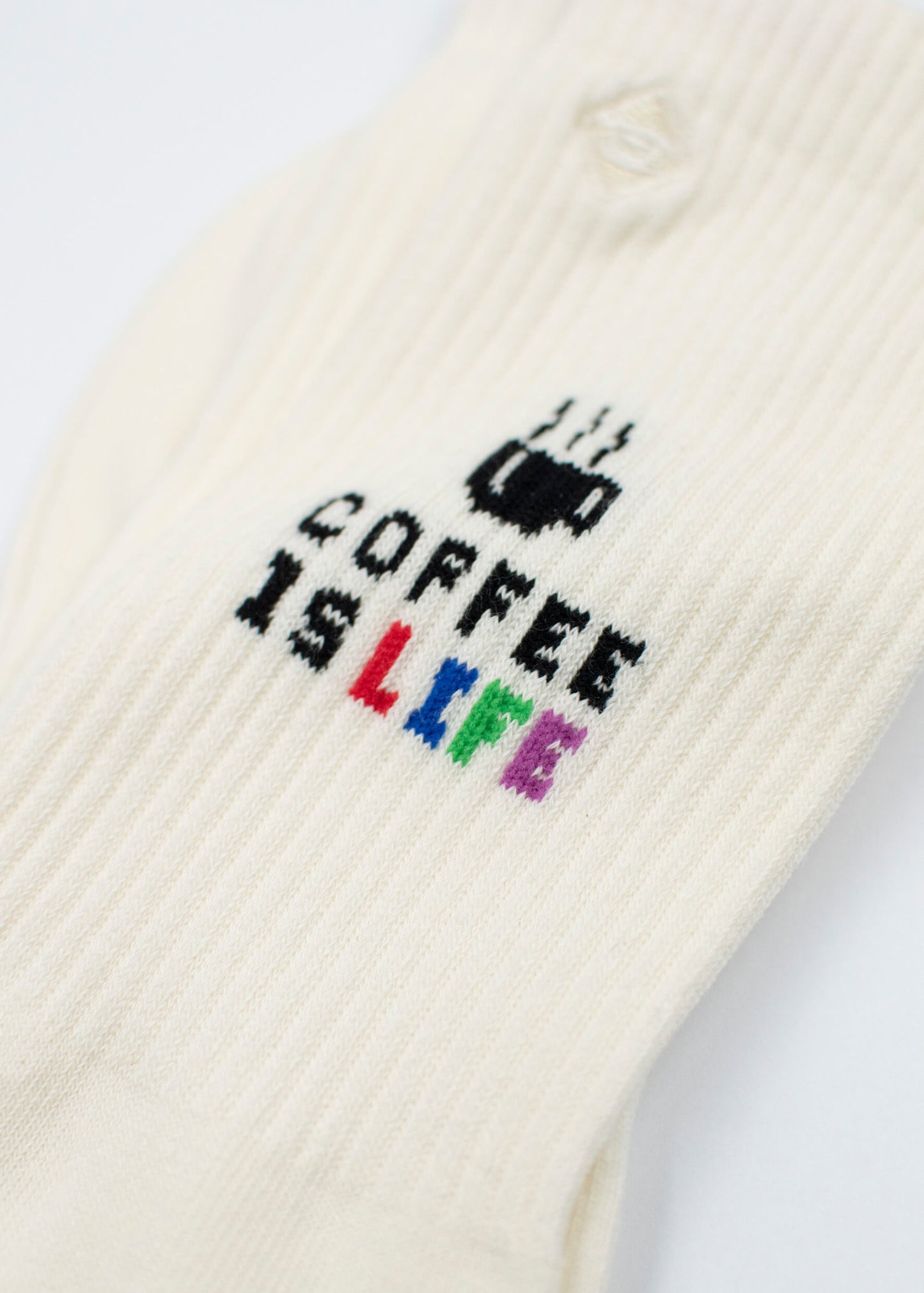 Eine Nahaufnahme von weißen gerippten J.Clay Tennissocken zeigt den Text COFFEE IS LIFE mit einer dampfenden Kaffeetasse. Das Wort LIFE hebt sich in Regenbogenfarben ab: rot, gelb, grün, blau und lila.
