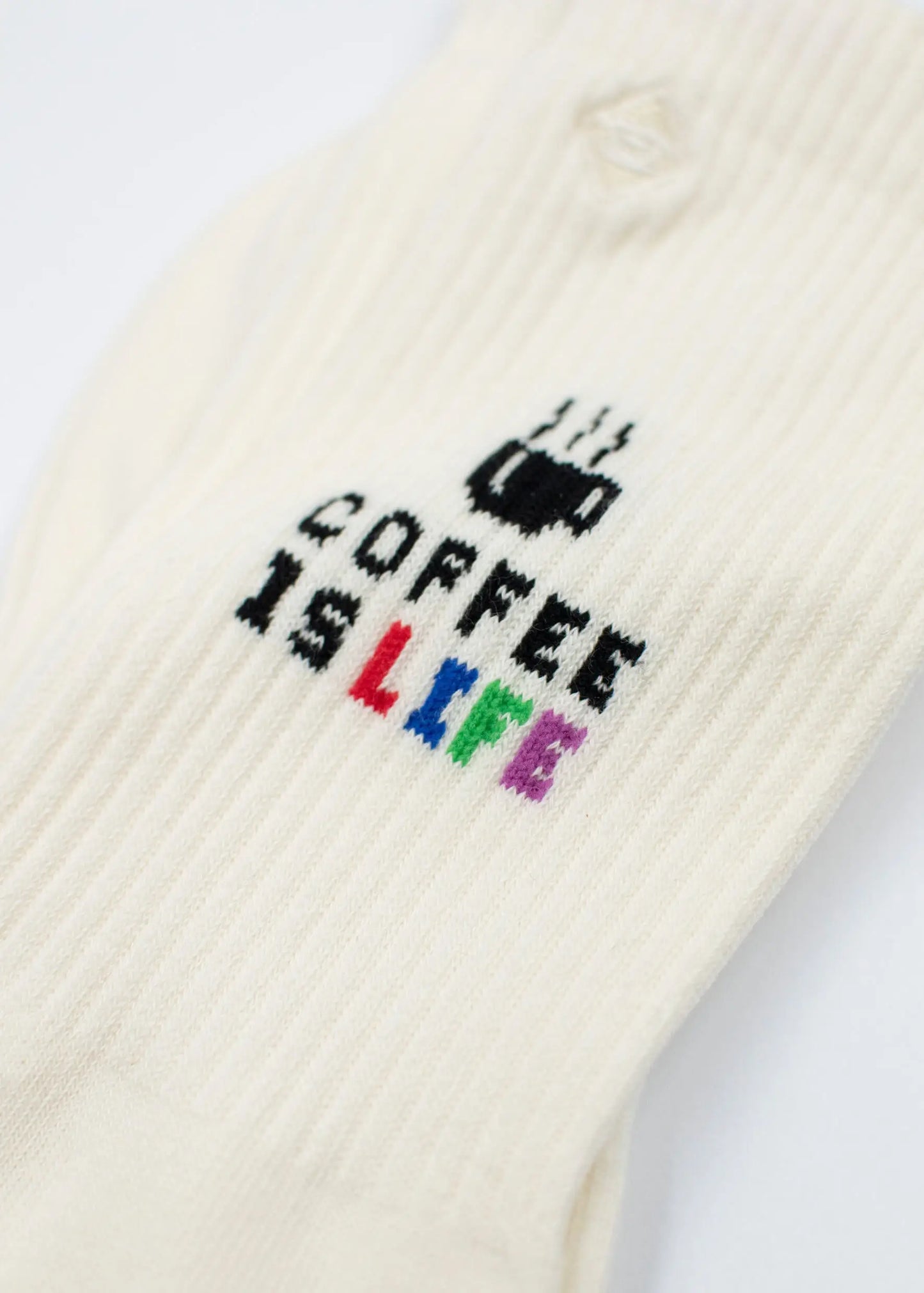 Eine Nahaufnahme von weißen gerippten J.Clay Tennissocken zeigt den Text COFFEE IS LIFE mit einer dampfenden Kaffeetasse. Das Wort LIFE hebt sich in Regenbogenfarben ab: rot, gelb, grün, blau und lila.