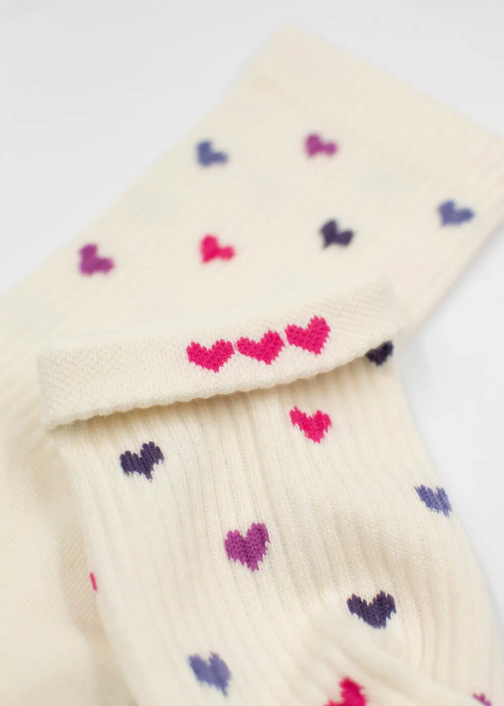 Eine Nahaufnahme der cremefarbenen J.Clay Tennissocken mit kleinen Herzmustern in Rosa, Lila und Blau. Die Socken liegen flach, ein Bündchen ist umgeschlagen und zeigt drei rosa Herzen in einer Reihe.