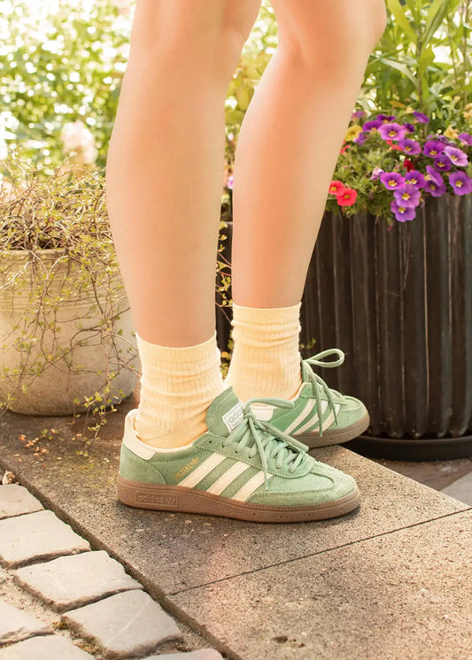 Eine Person in hellgelben J.Clay Damen Socken aus Bio Baumwolle und grünen Adidas-Turnschuhen steht auf einem Steinweg in der Nähe von Topfblumen und Grünpflanzen in hellem Sommerlicht.