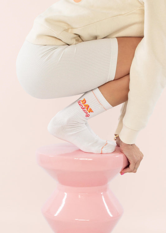 Eine Person in cremefarbenem Outfit und J.Clay Tennissocken mit orangefarbenem Text hockt auf einem glänzenden rosa Sockel vor einem hellrosa Hintergrund. Nur ein Teil des Körpers der Person ist zu sehen.