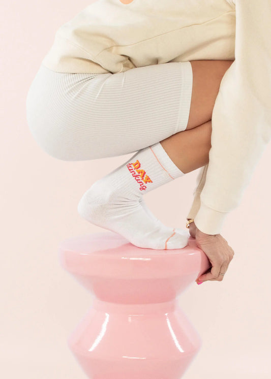 Eine Person in cremefarbenem Outfit und J.Clay Tennissocken mit orangefarbenem Text hockt auf einem glänzenden rosa Sockel vor einem hellrosa Hintergrund. Nur ein Teil des Körpers der Person ist zu sehen.