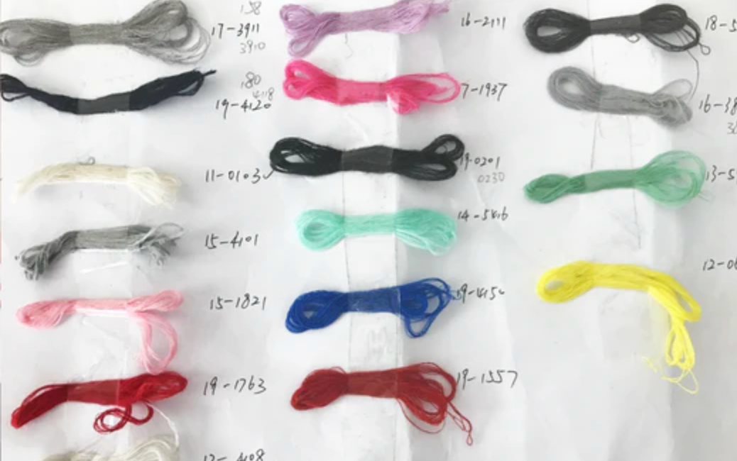 Dreizehn Bündel Stickgarn in verschiedenen Farben sind auf weißem Papier ausgelegt, jedes mit handgeschriebenen Nummern versehen. Die Farben sind schwarz, rosa, grau, weiß, mint, blau, gelb, rot und mehr.