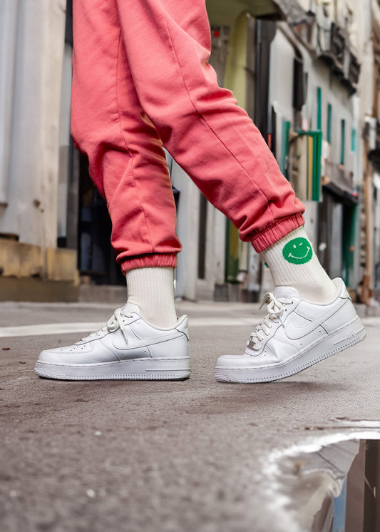 Eine Person läuft auf einer nassen Stadtstraße in rosa Jogginghosen, weißen Turnschuhen und J.Clay-Tennissocken mit einem grünen Smiley-Gesicht; nur ihre Unterschenkel und Füße sind in der Nähe einer Pfütze zu sehen.