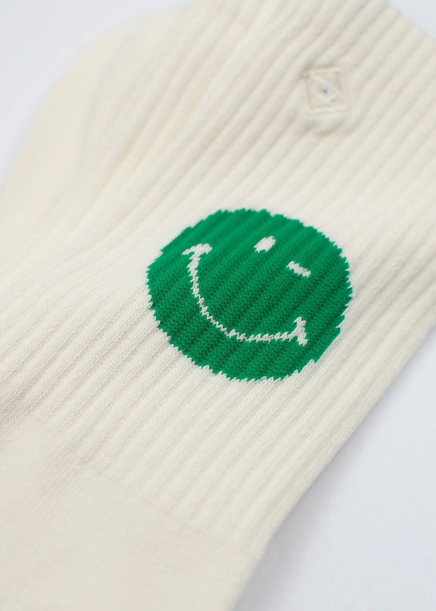 Nahaufnahme einer cremefarbenen gerippten Tennissocke von J.Clay mit einem aufgestickten grünen Smiley-Gesicht mit einem zwinkernden Auge in der Nähe des Knöchels.