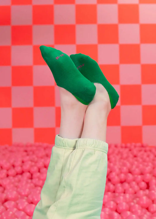 Eine Person, die leuchtend grüne J.Clay unsichtbare Füßlinge und helle Hosen trägt, hebt ihre Beine in die Luft. Rosa Bälle bedecken den Boden, während eine rot-rosa Schachbrettwand den Hintergrund bildet.