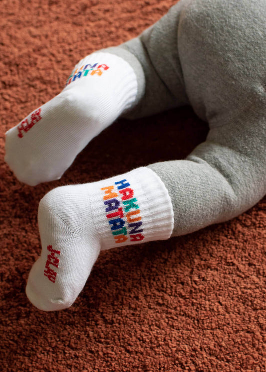 Ein Baby in grauer Hose liegt auf einem strukturierten braunen Teppich und trägt J.Clay Baby Socken - weiße Socken mit bunten HAKUNA MATATA Buchstaben. Ideal für Kinder Socken 0-4 Jahre Sammlungen.