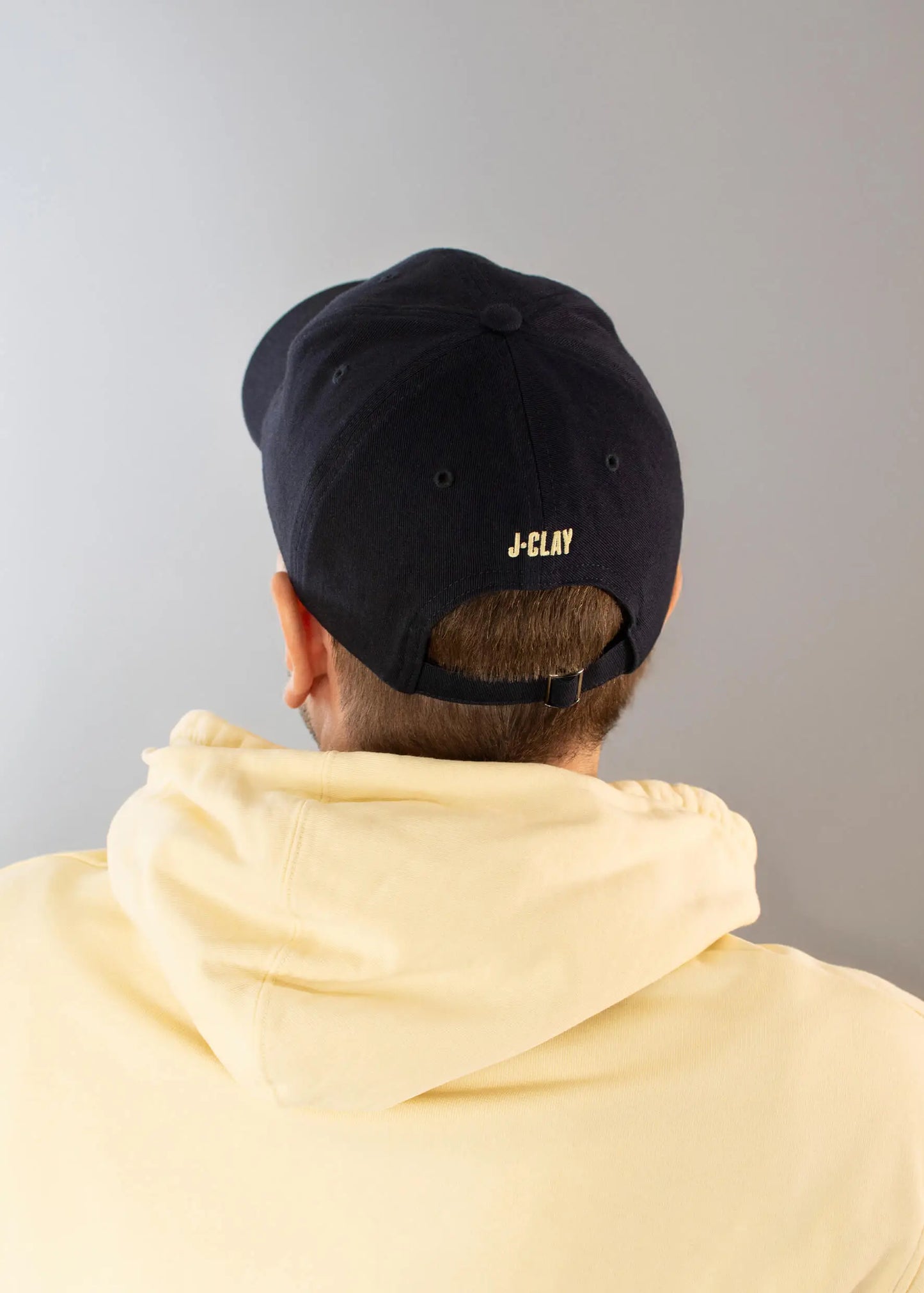 Eine Person, die einen hellgelben J.Clay-Kapuzenpullover und eine schwarze J.Clay-Vatermütze trägt, fotografiert von hinten vor einem einfarbig grauen Hintergrund.