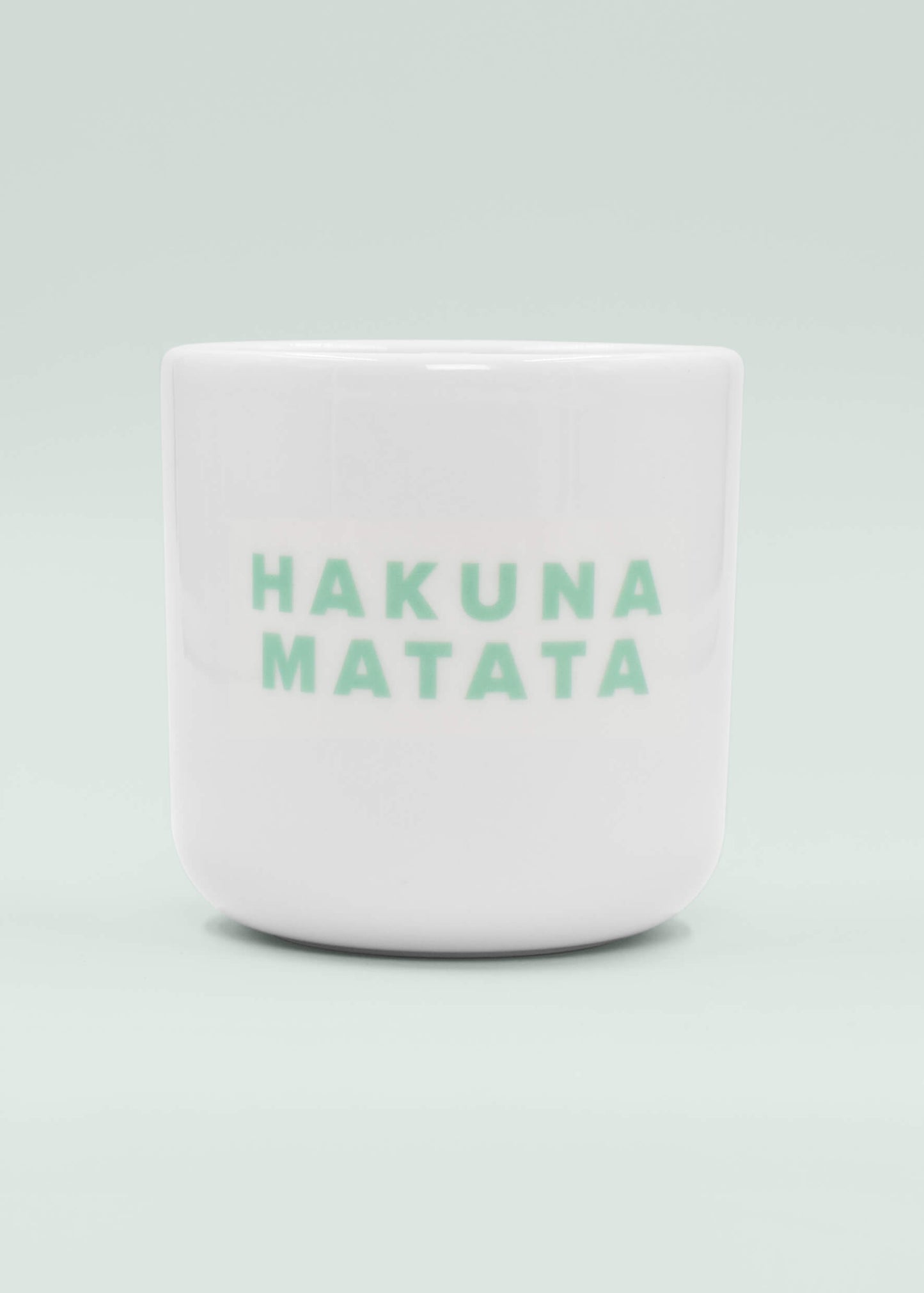 Eine weiße Keramik-Tasse ohne Henkel von J.Clay mit der Aufschrift "HAKUNA MATATA" in hellgrünen Großbuchstaben auf der Vorderseite vor einem hellen Hintergrund.