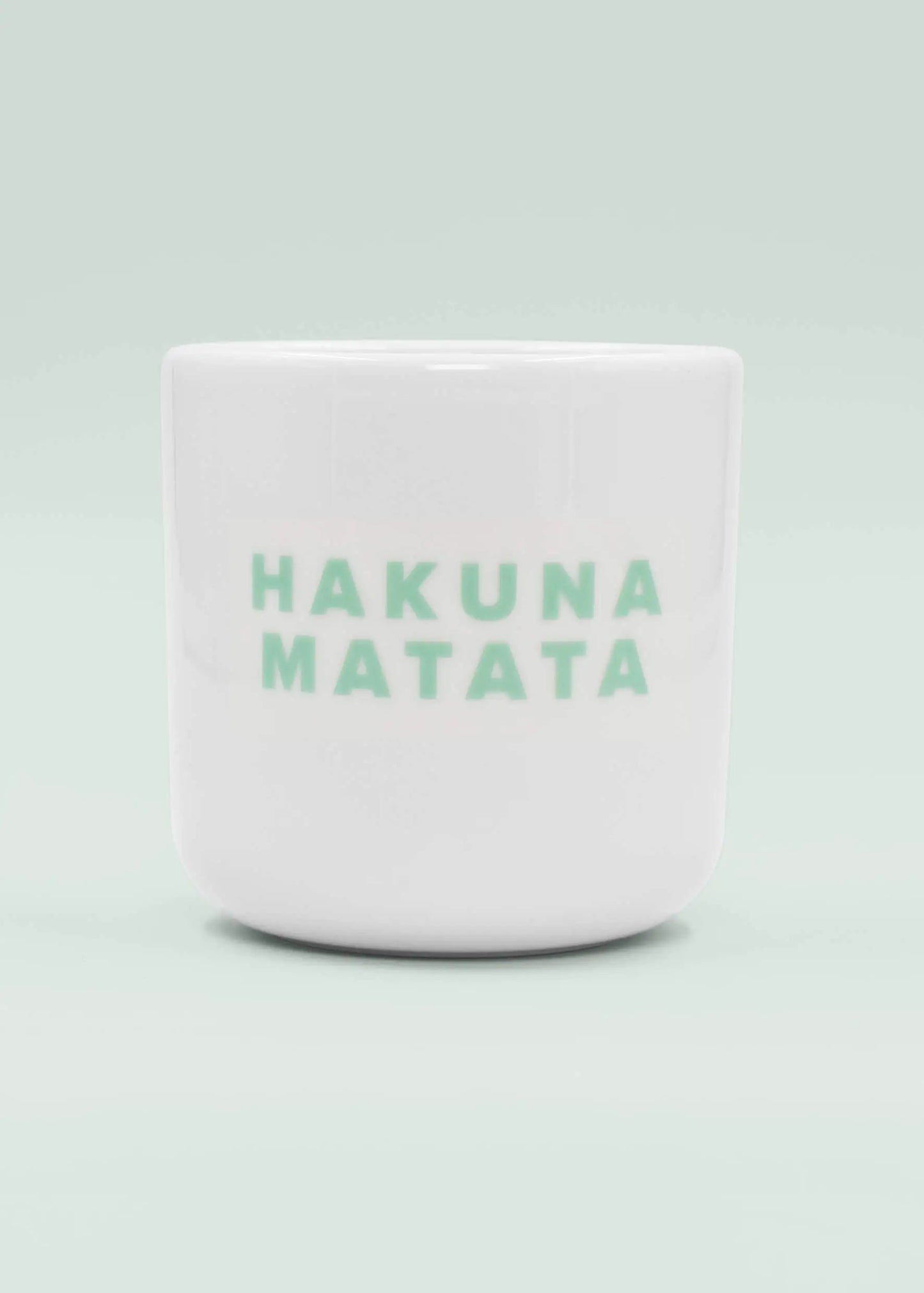 Eine weiße Keramik-Tasse ohne Henkel von J.Clay mit der Aufschrift "HAKUNA MATATA" in hellgrünen Großbuchstaben auf der Vorderseite vor einem hellen Hintergrund.