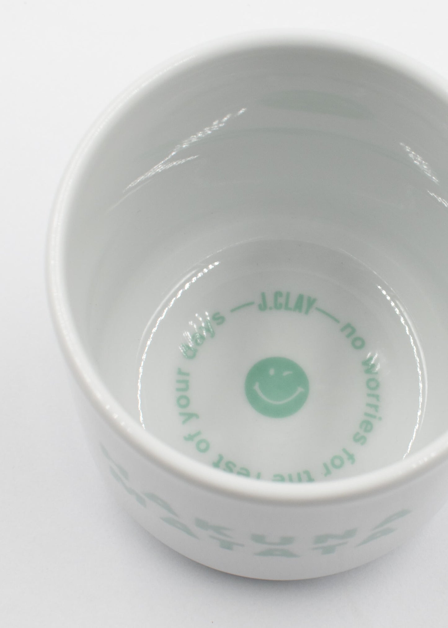 Eine weiße Keramikschale aus dem Hakuna-Matata-Paket von J.Clay trägt einen mintgrünen Text und ein Smiley-Gesicht im Inneren: "J.CLAY - no worries for the rest of your days" (J.CLAY - keine Sorgen für den Rest deiner Tage), abgebildet in einem Überkopf-Foto.