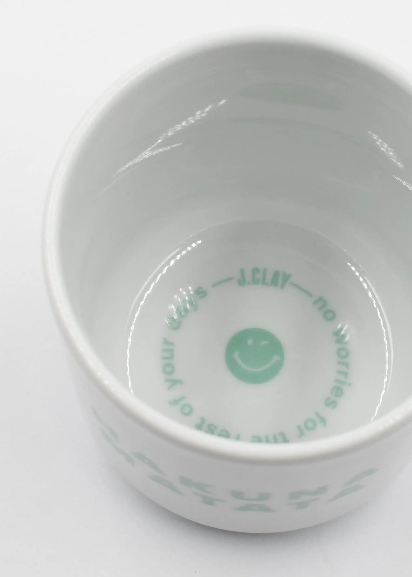 Eine weiße Keramikschale aus dem Hakuna-Matata-Paket von J.Clay trägt einen mintgrünen Text und ein Smiley-Gesicht im Inneren: "J.CLAY - no worries for the rest of your days" (J.CLAY - keine Sorgen für den Rest deiner Tage), abgebildet in einem Überkopf-Foto.