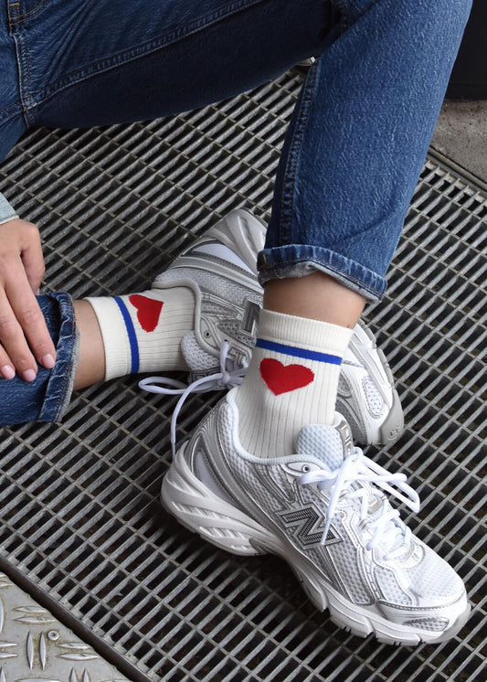 Eine Person in weißen Turnschuhen, blauen Jeans und J.Clay Red Heart Damen Socken - weiße Socken mit blauen Streifen und roten Herzen - sitzt auf einem Metallgitter, ein Bein gestreckt, das andere angewinkelt.