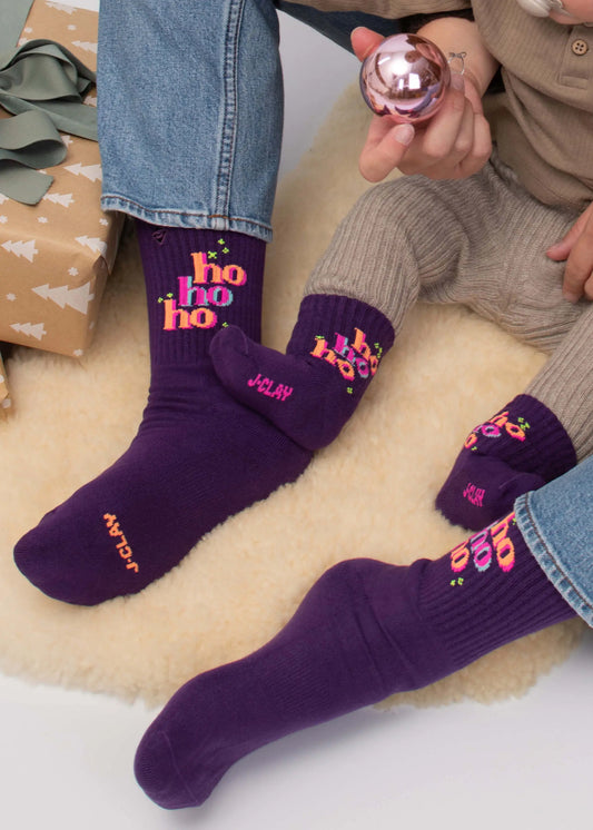 Zwei Kinder in „J.Clay ho ho ho“-Socken sitzen auf einem cremefarbenen Teppich neben verpackten Geschenken.