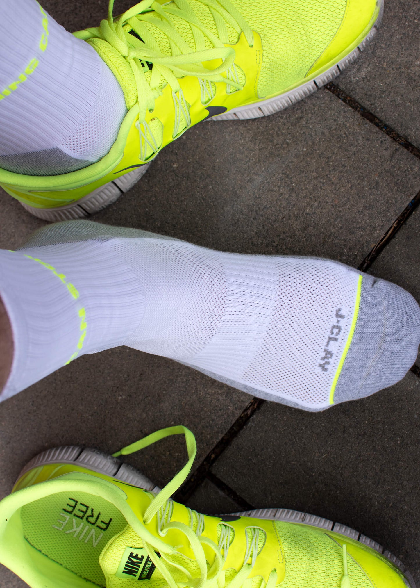 Eine Person in neongelben Nike Free Turnschuhen und atmungsaktiven weißen Sportsocken von J.Clay steht auf grauem Pflaster; ein Schuh ist ausgezogen und zeigt eine Socke mit gepolsterter Sohle, grauer Spitze und J.Clay-Schriftzug.
