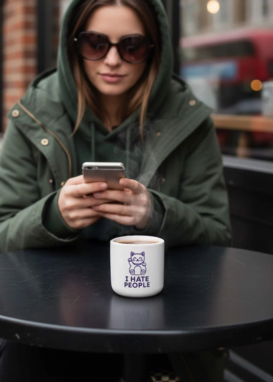 Frau mit Sonnenbrille schreibt eine SMS und hält eine J.Clay Hate Cat - Tasse mit "I HATE PEOPLE" und Katzenmotiv.