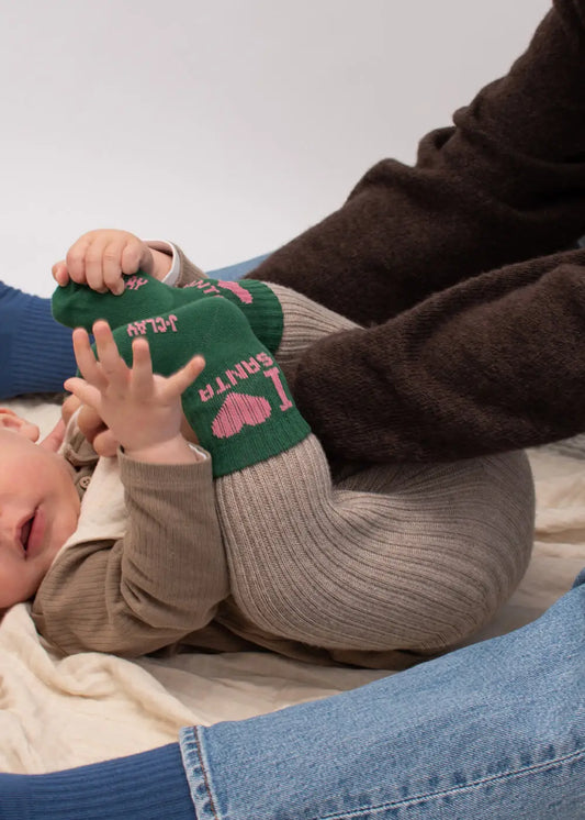 Baby auf einer Decke in „I Love Santa – Mini“ von J.Clay, beigefarbene Kleidung, grüne Socken, spielt mit den Füßen.