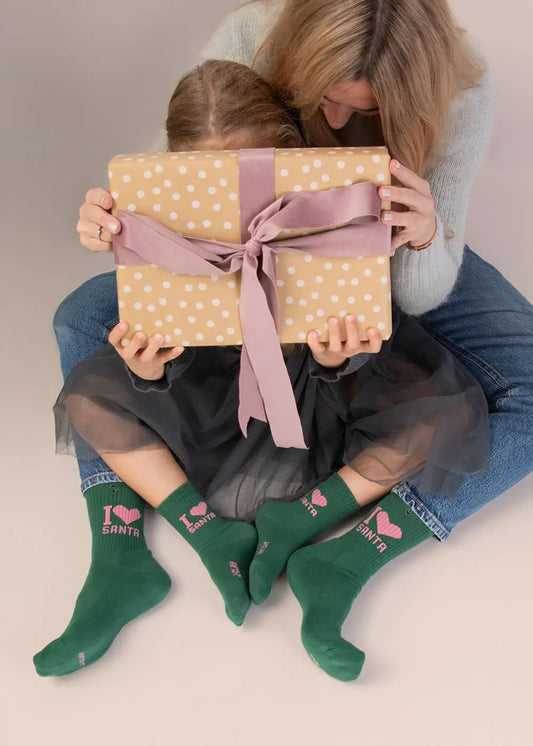 Frau und Kind mit einem gepunkteten Geschenk, die grüne Kindersocken von J.Clay mit der Aufschrift „I Love Santa“ tragen.