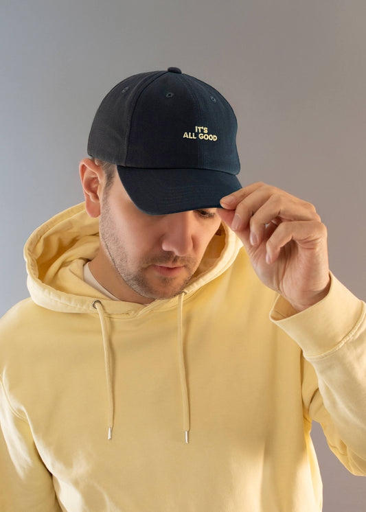 Ein Mann in einem hellgelben J.Clay-Kapuzenpulli und einer dunklen J.Clay-Vatermütze mit der Aufschrift "ITS ALL GOOD" verstellt mit einer Hand die Krempe seiner Mütze vor einem unifarbenen Hintergrund.