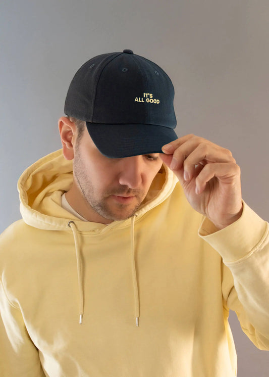 Ein Mann in einem hellgelben J.Clay-Kapuzenpulli und einer dunklen J.Clay-Vatermütze mit der Aufschrift "ITS ALL GOOD" verstellt mit einer Hand die Krempe seiner Mütze vor einem unifarbenen Hintergrund.