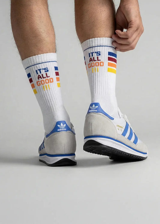 Eine Person rockt Streetstyle in den All Good-Socken von J.Clay - weiß mit bunten Streifen und "ITS ALL GOOD", gepaart mit hellgrauen Adidas-Sneakern mit blauen Streifen, die auf einem passenden grauen Boden gute Laune verbreiten.
