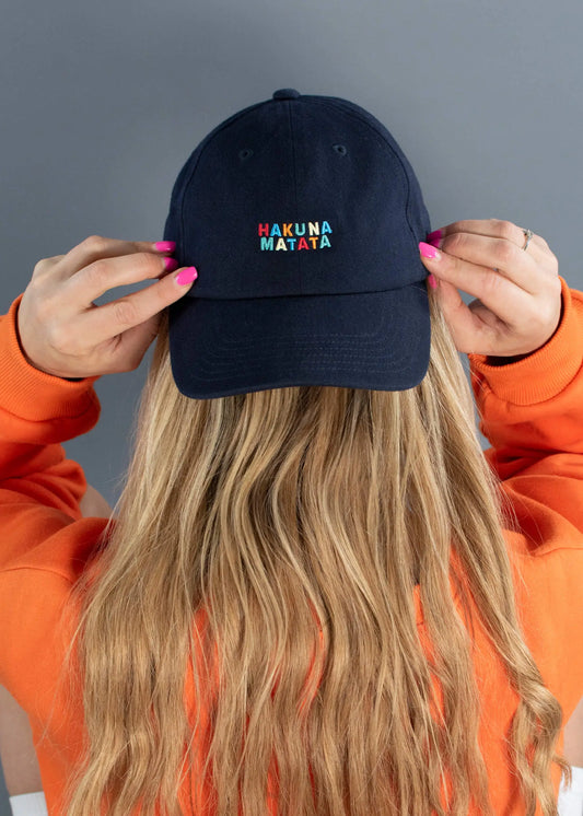 Eine Person mit langen blonden Haaren in einem orangefarbenen Sweatshirt rückt eine marineblaue J.Clay Dad Cap zurecht, auf der in bunten Buchstaben "HAKUNA MATATA" vor einem schlichten grauen Hintergrund aufgestickt ist.