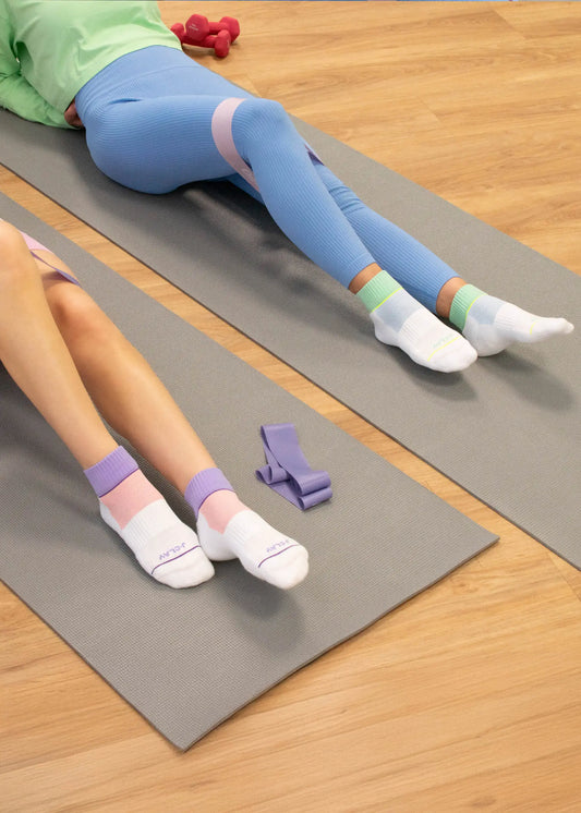 Zwei Personen tragen J.Clay Sportsocken mit gepolsterter Sohle und liegen in bunten Leggings und weißen Socken mit pastellfarbenen Bändern auf grauen Yogamatten. Ein Widerstandsband und eine Hantel liegen in der Nähe auf dem Holzboden und zeigen nur ihre Beine und Füße.