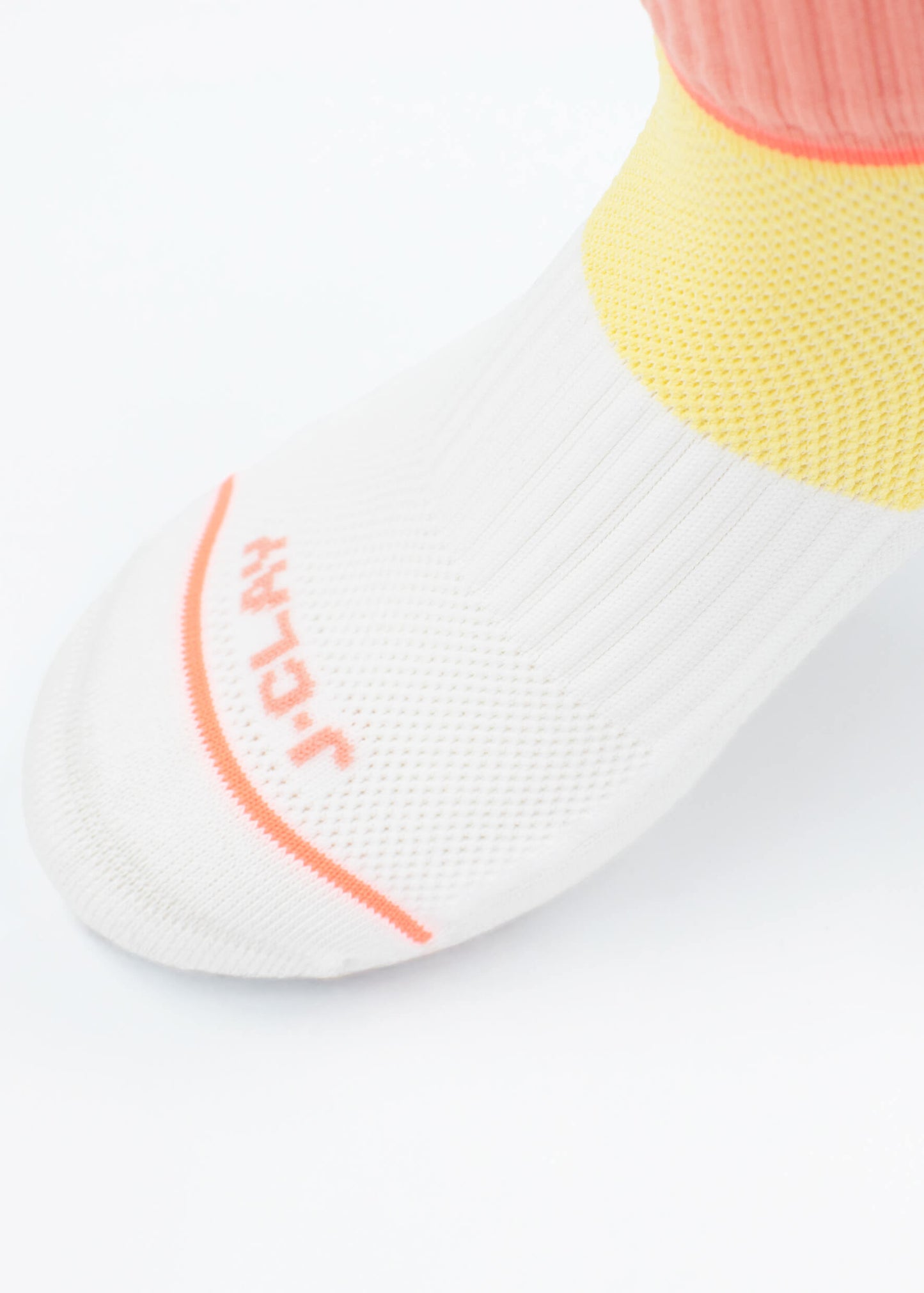 Eine Nahaufnahme eines Fußes in einer weißen J.Clay Sportsocke mit orangefarbenen Akzenten, "JCLAY" in der Nähe der Zehen und gerippten gelben und rosafarbenen Details um den Knöchel, aus atmungsaktivem Material für Komfort.