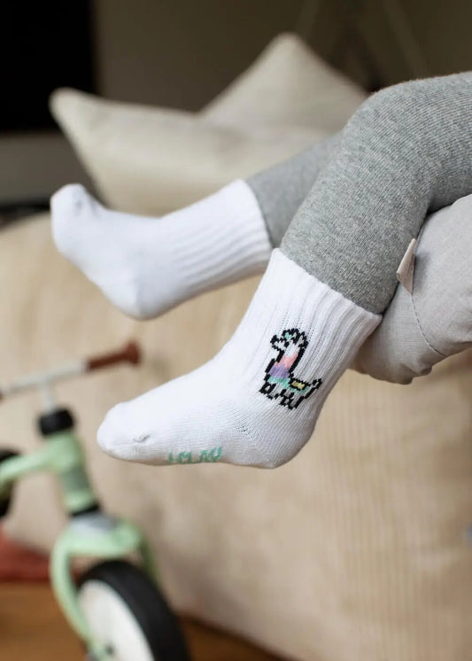 Ein Kind in grauen Leggings und J.Clay bunte Baby Socken mit einem bunten Dinosauriermotiv sitzt auf einer beigen Couch, während im Hintergrund ein grünes Dreirad zu sehen ist.