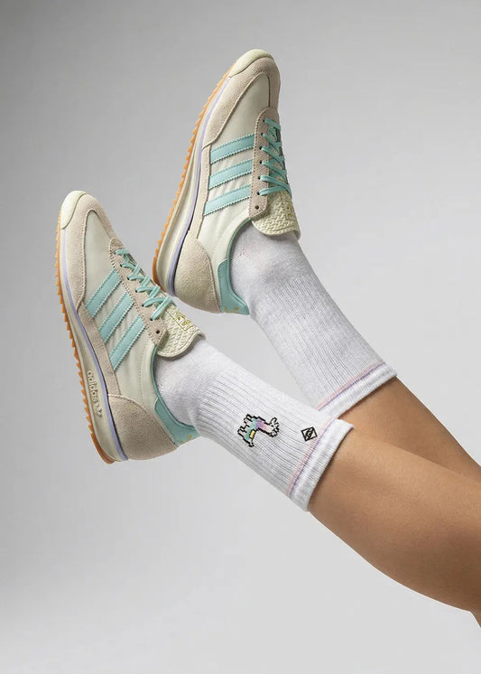 Eine Person mit heller Haut trägt J.Clay Lama Tennissocken und cremefarbene Adidas-Turnschuhe mit hellblauen Streifen und Gummisohlen, die Beine vor einem einfarbig grauen Hintergrund.