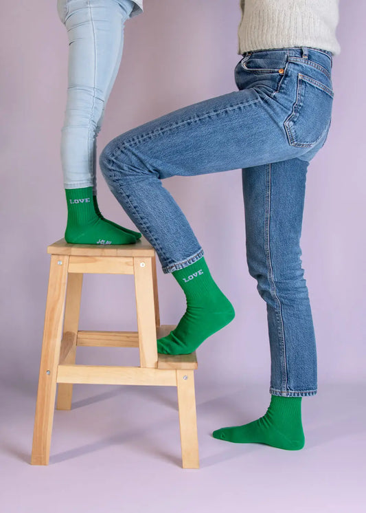 Zwei Kinder in J.Clay Love-Socken und Jeans stehen auf einem Hocker vor einem hellvioletten Hintergrund.