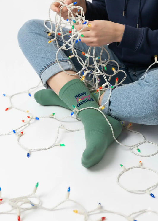 Person in grünen "Merry Christmas"-Socken, die eine verworrene Kette von bunten Lichtern hält.