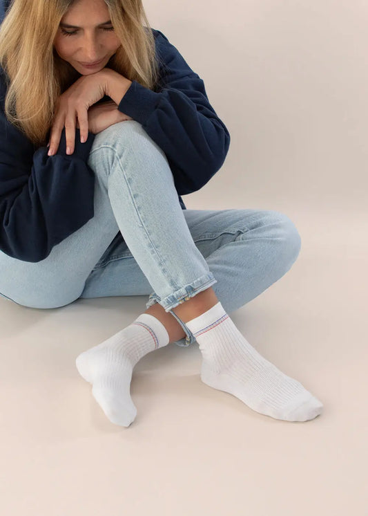 Eine Frau mit langen blonden Haaren sitzt auf dem Boden, die Knie hochgezogen, und trägt einen marineblauen Pullover, hellblaue Jeans und J.Clay mittelhohe Damensocken aus Bio-Baumwolle. Sie blickt sanft nach unten. Marke: J.Clay.