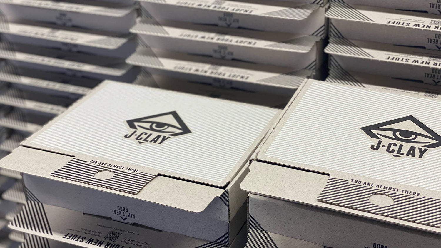 Stapel von weißen Pizzakartons mit dem J-CLAY Logo, das ein stilisiertes Auge in einem Dreieck zeigt. Die Kartons sind ordentlich angeordnet, und auf der vorderen Klappe ist der Satz YOU ARE ALMOST THERE aufgedruckt.