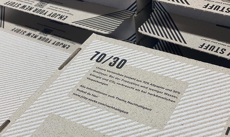 Eine Nahaufnahme einer umweltfreundlichen Kartonverpackung mit der Aufschrift 70/30 und einer Erläuterung auf Deutsch, dass der Karton zu 70 % aus Recyclingpapier und zu 30 % aus Grasfasern besteht. Weitere Informationen zur Nachhaltigkeit finden Sie weiter unten.
