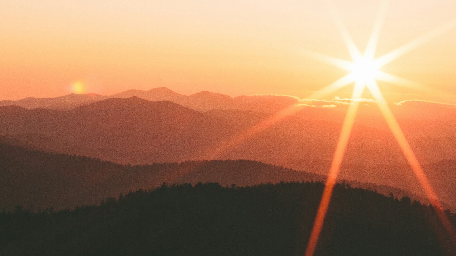 Der Sonnenaufgang über einer Schicht von Gebirgskämmen. Die Sonne scheint hell und wirft warme orangefarbene und goldene Farbtöne über den Himmel und die Landschaft.