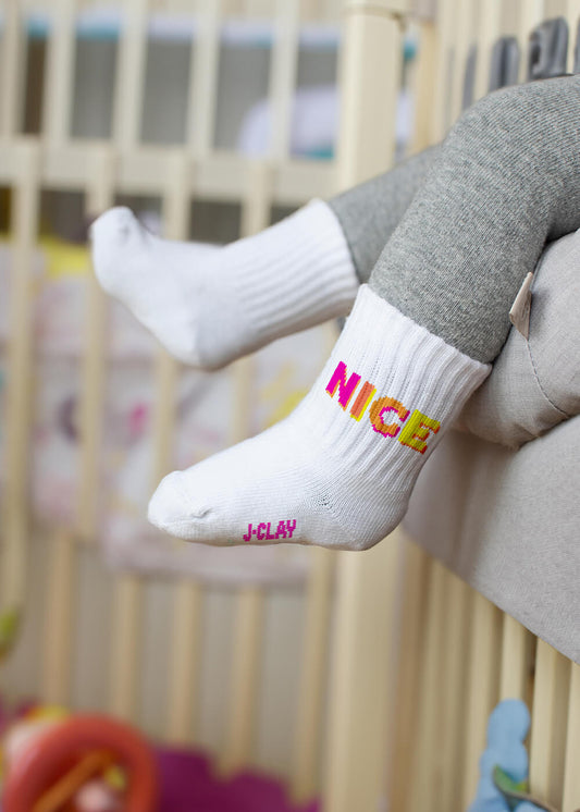 Ein Kind in grauer Hose sitzt auf einem hellen Sofa und trägt J.Clay Baby Socken: auf einer steht "NICE" in gelb/rosa, auf der anderen "J-CLAY" in rosa. Ideal für Kinder im Alter von 0-4 Jahren. Im Hintergrund sind ein Kinderbett und Spielzeug zu sehen.