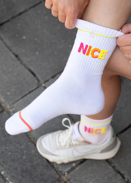 Eine Person in weißen Turnschuhen und J.Clay-Tennissocken mit leuchtend pinkem und gelbem "NICE"-Schriftzug sitzt auf grauem Steinpflaster und passt eine Socke an.