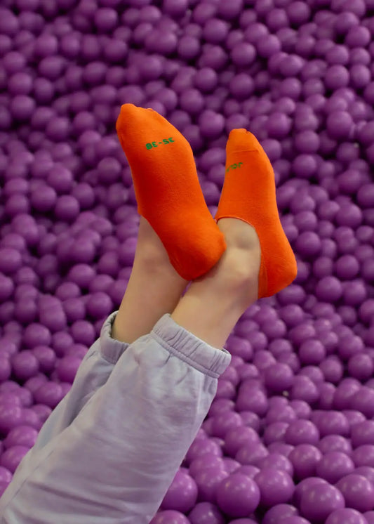 Eine Person, die leuchtend orangefarbene J.Clay-Socken und eine helllavendelfarbene Hose trägt, hebt ihre Beine über eine mit lila Plastikbällen gefüllte Ballgrube, selbstbewusst, als hätte sie rutschfeste Füßlinge an.