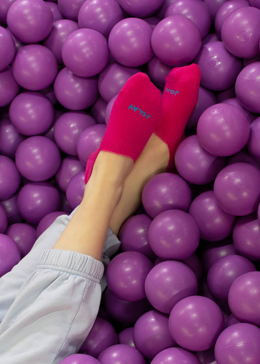 Eine Person, die leuchtend pinke J.Clay-Socken und eine hellblaue Hose trägt, ruht ihre Füße auf einem Stapel lila Plastikbälle aus und genießt den Komfort der rutschfesten Füßlinge.