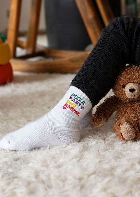 Ein Kind trägt J.Clay Baby Socken (PIZZA, PARTY, UNO & MORE) und sitzt mit einem braunen Teddybären auf einem flauschigen Teppich. Nur die Beine und das Spielzeug sind zu sehen - ideal für J.Clay Kinder Socken von 0-4 Jahren.
